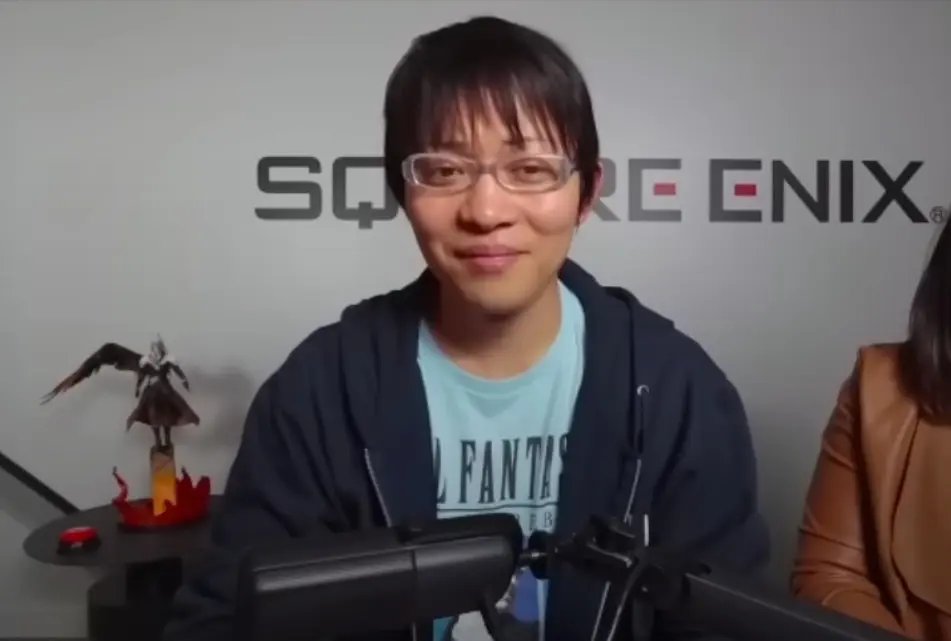Naoki Hamaguchi i glasögon och blå t-shirt, sittandes framför en Square Enix-vägg.