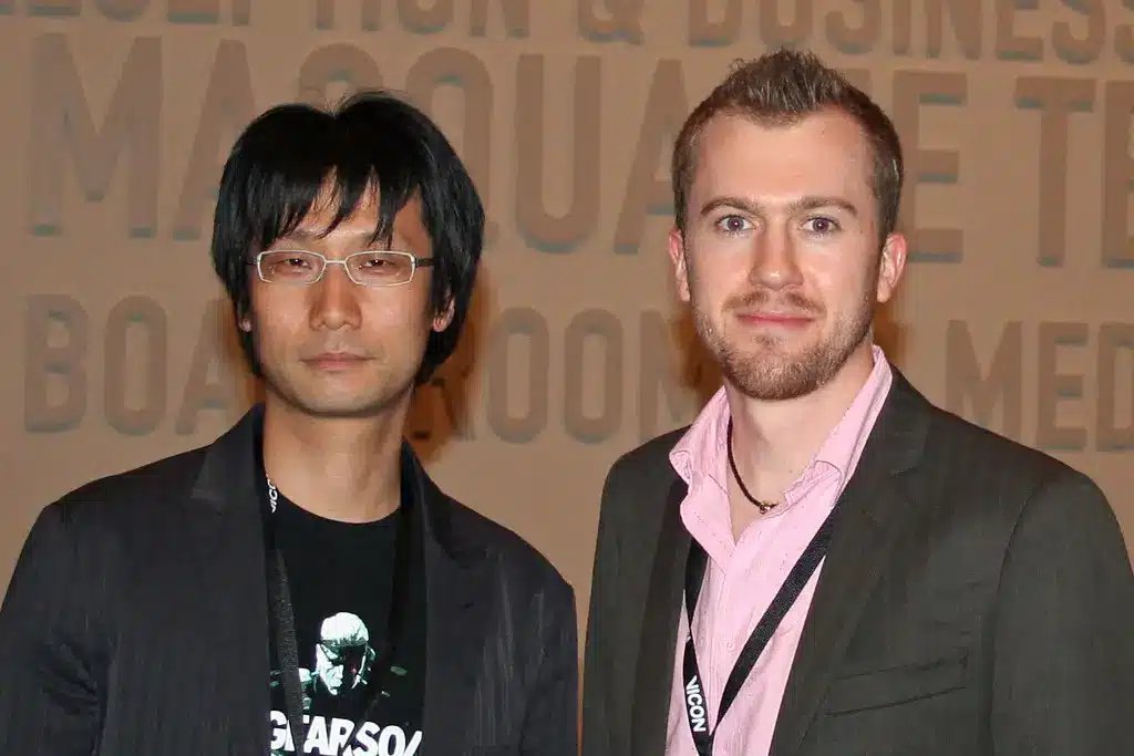 Hideo Kojima i glasögon och svart kavaj, stående bredvid en annan man.