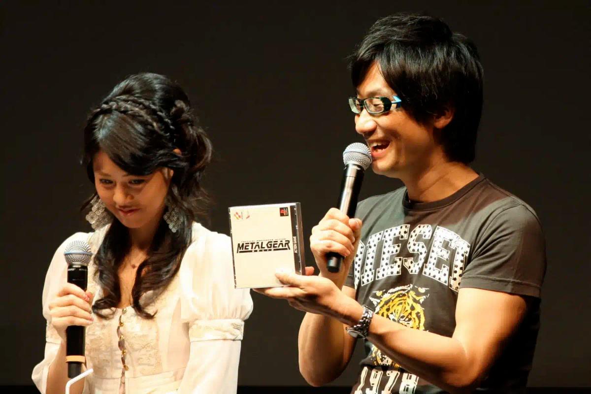 Hideo Kojima håller i ett Metal Gear-fodral samtidigt som han talar i en mikrofon.